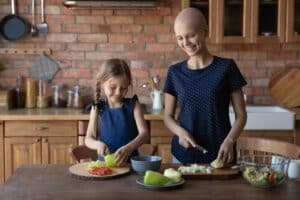 person undergoing chemotherapy smiling while cutting vegetables with a young child in a kitchen
